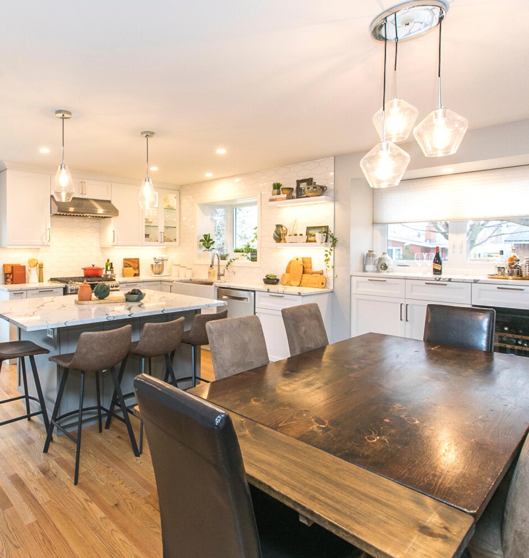 Shot of kitchen from dining area overlooking solid wood table, leather chairs and a bright white newly-renovated kitchen